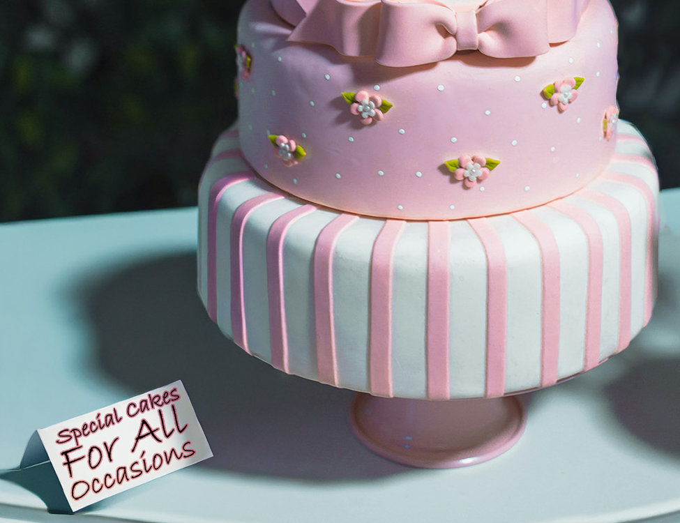 On a table there is a two tiered birthday cake with pink and white striped icing and flowers. There is an icing bow accross the top. There is a card on the table that reads ''Special Cakes For All Occasions'
